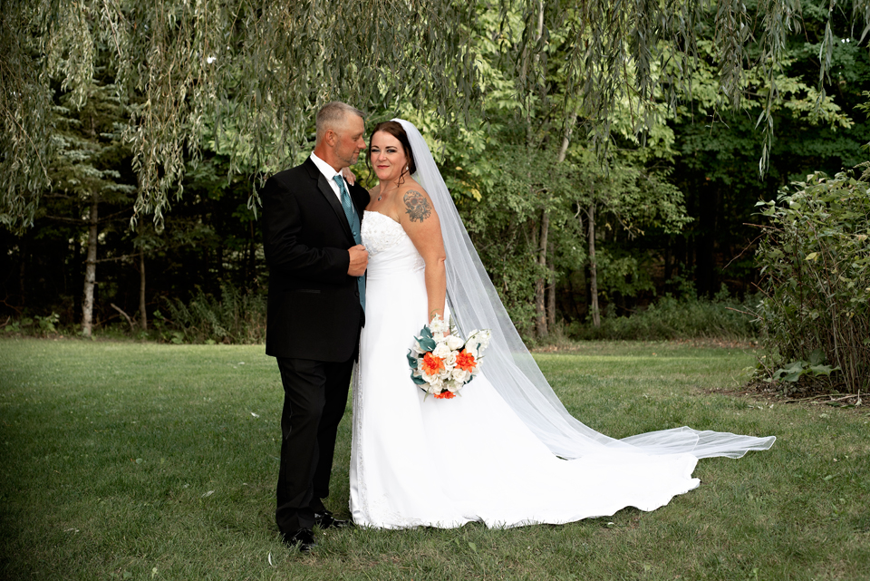 Bride and Groom standing together in front of weeping willow trees in Rose City, MI