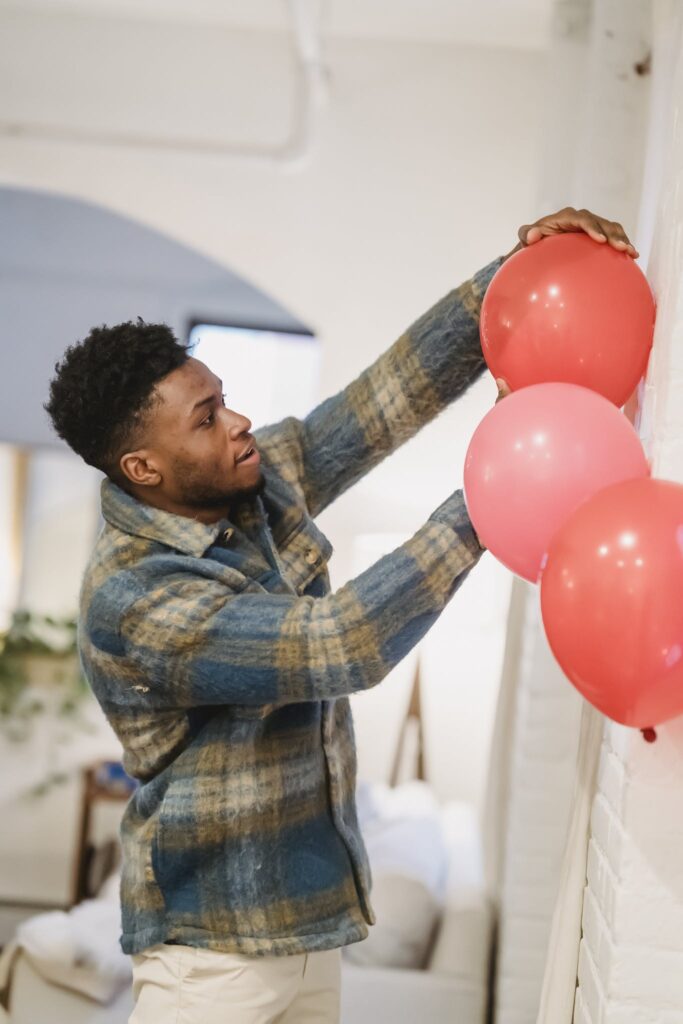 Side view of young bearded African American male in casual clothes standing near white wall and decorating room with inflatable balloons
