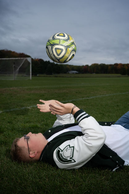 High School senior boy laying on his back on the ground throwing a soccer ball up in the air.