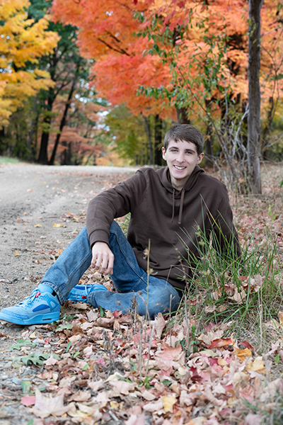 High School Senior Boy sitting in leaves with colors of fall in background