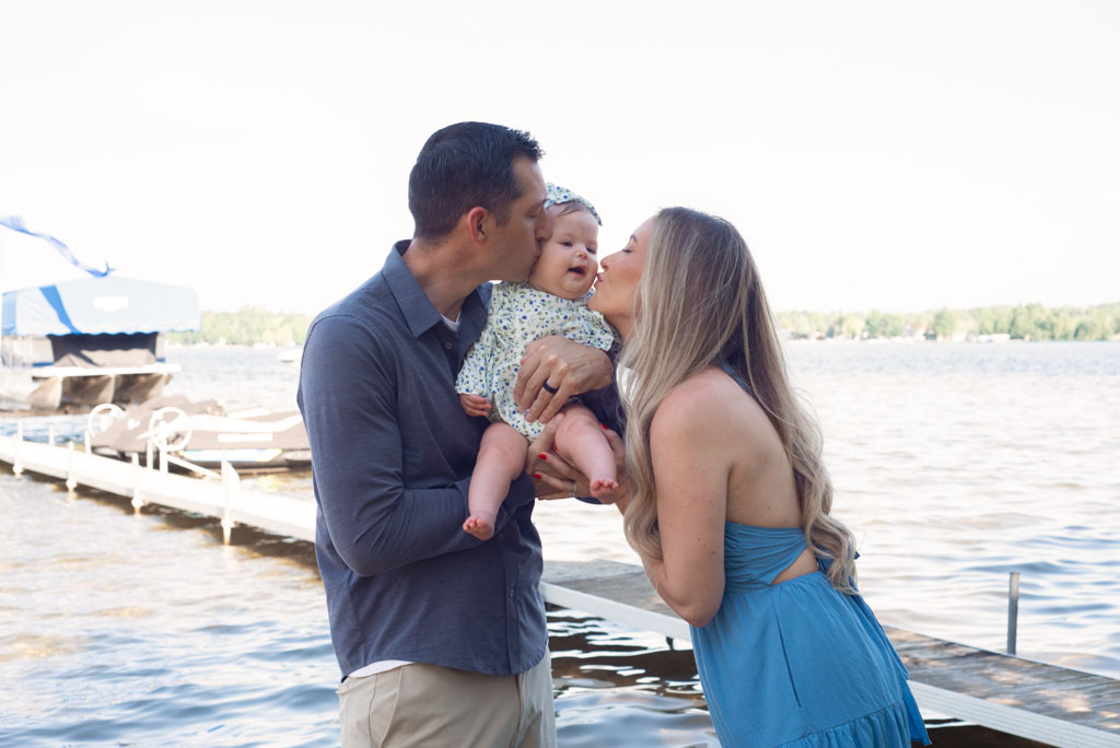 Mom and dad holding baby between them and kissing it on the cheek.
