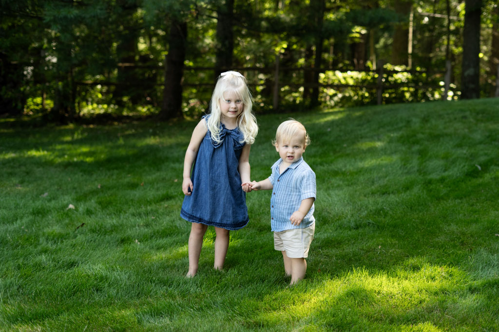 Big sister and little brother holding hands in a grassy backyard.